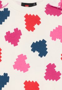 Tela blanca con un patrón de corazones pixelados en rosa, rojo y azul oscuro. Presenta un cuello redondo y pequeños acentos rosas en los hombros.