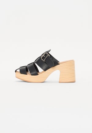 Black leather clog-style mules with a wooden block heel, featuring crisscross straps and a metal buckled strap for adjustment.