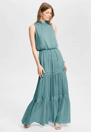 Femme portant une robe maxi sans manches couleur sarcelle avec taille froncée, debout, regardant vers le bas, posant devant un fond clair uni.