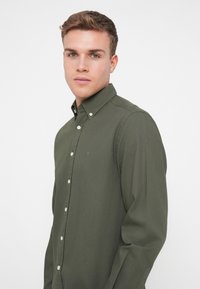 Camisa de botonadura en verde oscuro con cuello, mangas largas y corte ajustado. Presenta botones blancos y un logo sutil en el pecho.