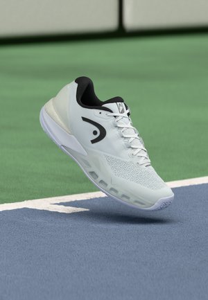 Valkoinen urheilukenkä, jossa musta logo ja pohjan yksityiskohdat, leijuu sinivihreän tenniskentän pinnan yläpuolella.