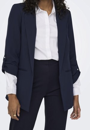 Blazer bleu marine avec une coupe ajustée, tissu léger et poignets roulés. Associé à une chemise blanche bien ajustée, créant un look professionnel.