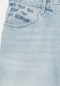 Light blue denim fabric with dark blue embroidered text above a front pocket reading, "WRITE YOUR OWN SCRIPT THEN LIVE ACCORDING."
