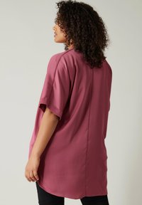 Blusa magenta con maniche corte, texture setosa, vestibilità ampia e orlo arrotondato. Il dettaglio della cucitura sul retro aggiunge un interesse sottile al design.