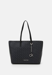 Sac fourre-tout noir Calvin Klein avec motif en all-over "ck", deux poignées, et un pendentif en argent en forme de "ck" sur fond blanc.