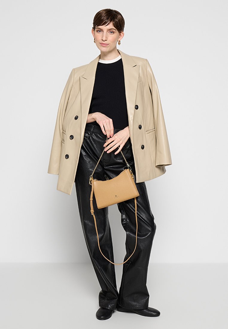 Bolso de mano de cuero color marrón claro con un acabado texturizado, que presenta una correa desmontable. El modelo lleva un blazer beige y pantalones de cuero negros.