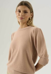 Mladá žena s blond vlasy má na sobě světle růžový vlněný svetr s nafouklými rukávy a zlaté kruhové náušnice, pózuje na jednobarevném pozadí.