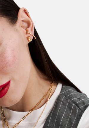 Goldener Creolen-Ohrring mit kleinen dekorativen Anhängern und glänzendem Finish. Das Modell trägt geschichtete goldene Halsketten zu einem gestreiften grauen Oberteil.