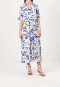 Květinové midi šaty v krémové barvě s modrými vzory, krátké rukávy, zapínání na knoflíky vpředu a zavázaný pas. Vysoké béžové sandály na tenkých řemíncích.