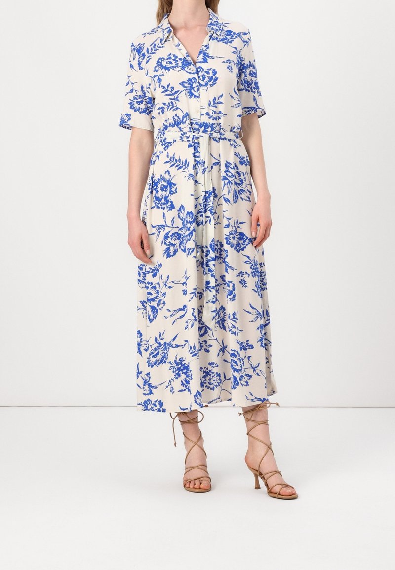 Květinové midi šaty v krémové barvě s modrými vzory, krátké rukávy, zapínání na knoflíky vpředu a zavázaný pas. Vysoké béžové sandály na tenkých řemíncích.