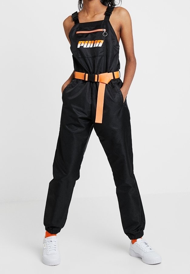 Femme portant une combinaison noire Puma avec une ceinture orange et une fermeture éclair, associée à des baskets blanches et des chaussettes orange, debout les mains dans les poches.
