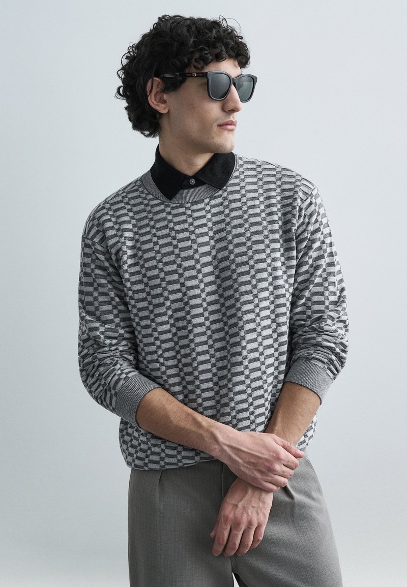 Camisola cinza com padrão e gola preta, combinada com calças cinzentas. Modelo usa óculos de sol pretos, exibindo uma pose descontraída contra um fundo claro.