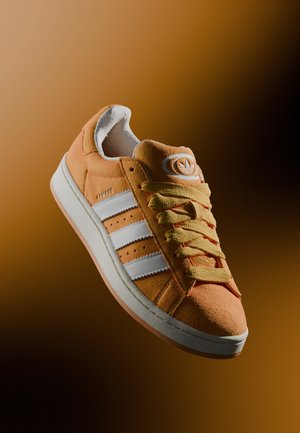 Orange ruskind Adidas sneaker med hvide striber og gule snørebånd, fremvist mod en gradient brun baggrund.