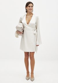 Robe enveloppante crème avec des manches longues, décolleté en V profond, lien sur le côté, et texture satin lisse. Le modèle tient une pochette beige assortie et porte des talons beiges.