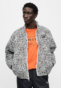 Μαύρο-λευκό μπουφάν με μοτίβο, ψηλό γιακά και κλείσιμο με φερμουάρ, φορεμένο πάνω από πορτοκαλί μπλουζάκι με μαύρο λογότυπο.