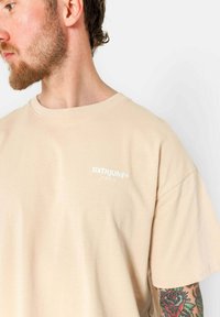 Beige katoenen t-shirt met een ronde halslijn en korte mouwen, voorzien van een wit "SIXTHUNE PARIS" logo op de borst. Glad textuur.