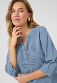 Blouse bleu clair, boutonnée, présentant des plis verticaux à l'avant et des manches trois-quarts avec des accents de boutons. Tissu doux et texturé.