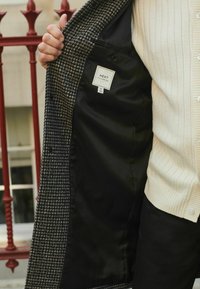 Palton cu model houndstooth, realizat dintr-un material texturat, cu interior din satin negru și etichetă care indică "NEXT TAILORING", mărimea M.