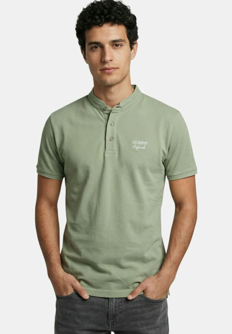 Homme portant un polo henley à manches courtes vert clair avec le logo "Lee Cooper Originals", debout les mains dans les poches d'un jean gris foncé.