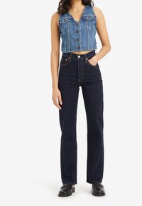 Femme portant un gilet en jean bleu sans manches boutonné, un jean taille haute bleu foncé et des chaussures noires, debout avec une main dans la poche.