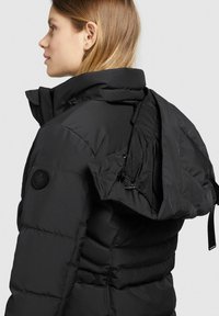 Sort pufferjakke med høj krave, lynlåset og snoretræk i hætten samt et logo-patch på ærmet. Glat, isoleret overflade.
