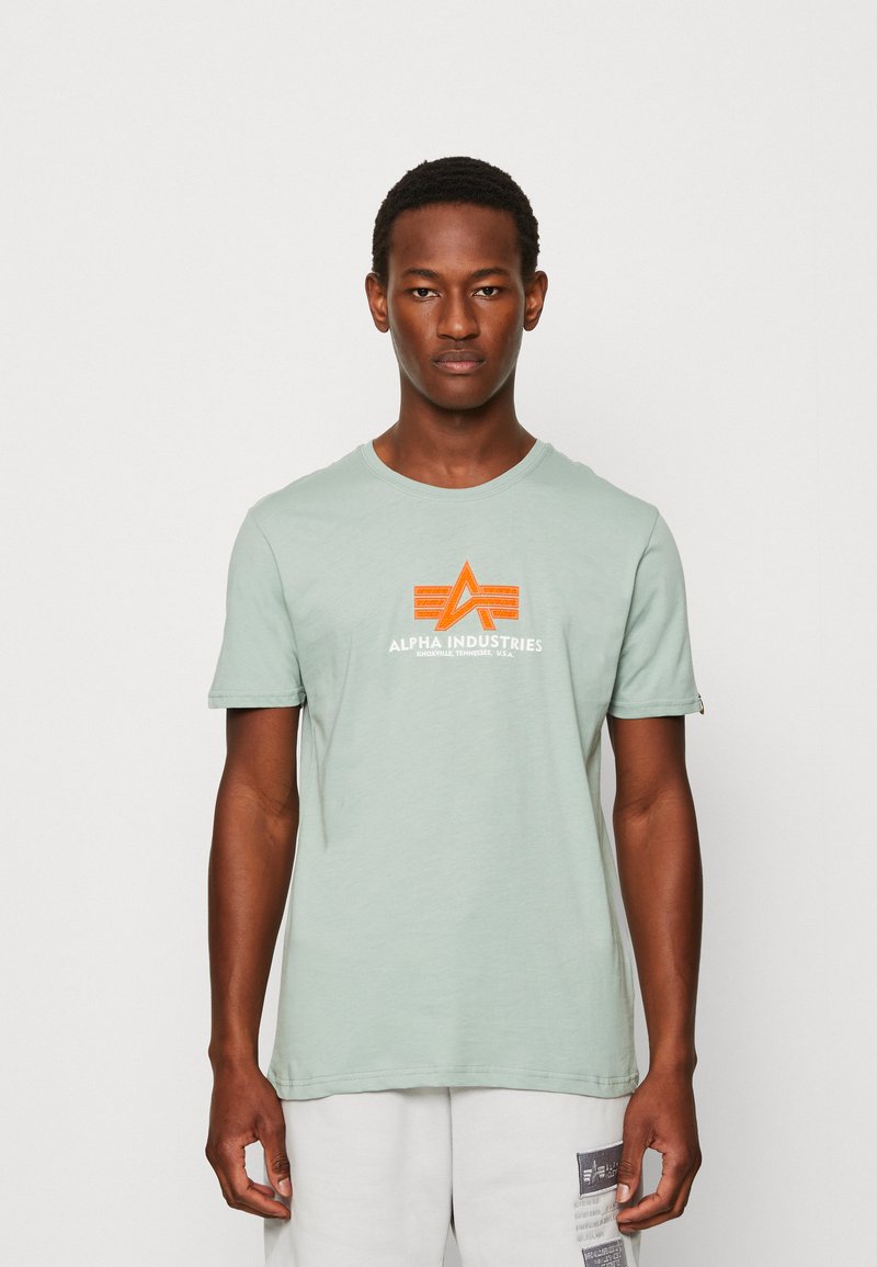Šviesiai žalia medvilninė marškinėliai su oranžiniu „ALPHA INDUSTRIES“ logotipu ir grafikos akcentais. Apvalus kaklas, standartinė apdirbimo forma, trumpi rankoviai.