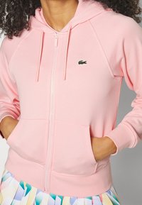 Žena v světle růžové mikině na zip s kapsami a malým zeleným logo krokodýla na hrudi, doplněná barevnou skládací sukní.