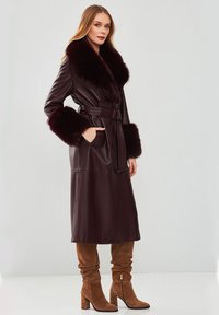 Cappotto in pelle bordeaux con colletto e polsini in pelliccia sintetica, vita con cintura, bottoni frontali e tasche laterali. Texture liscia, design lungo fino al pavimento.