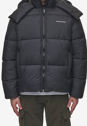 Mand iført en sort dunjakke med lynlås foran og hætte, kombineret med olivengrønne cargobukser.