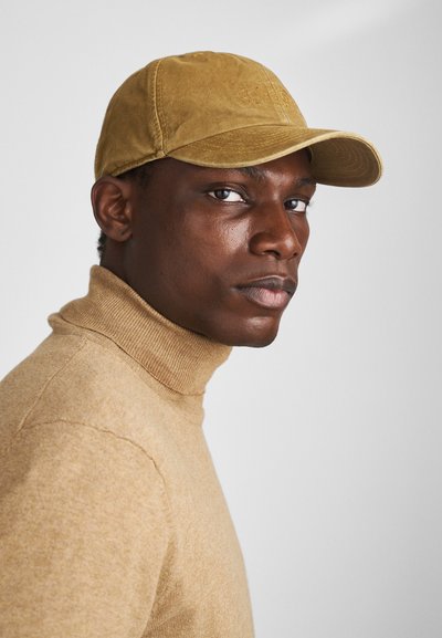 Κασετίνα μπέιζμπολ σε καφέ απόχρωση με καμπύλο γείσο, υφή και ανάγλυφο λογότυπο. Φοριέται με εκρού ζιβάγκο πουλόβερ, με ραμμένο λαιμό.