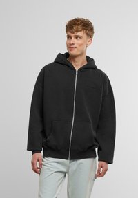 Črna zip-up hoodie iz mehkega materiala, ki ima velik sprednji žep, kapuco z vrvico in rebraste manšete. Minimalno oblikovanje blagovne znamke na prsih.