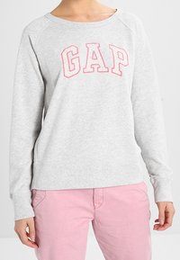 Felpa grigia con maniche raglan e colletto a girocollo, che presenta la parola "GAP" in lettere rosa sul petto. Tessuto morbido.