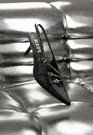 Svart slingback-sko med spetsig tå, Krokodilstruktur, guldnitar och spännbanddetaljer, mot en skinande silverbakgrund.