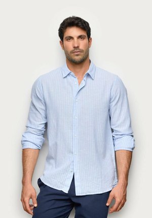 Mężczyzna z krótkimi ciemnymi włosami, ubrany w jasnoniebieską, paskowaną koszulę z podwiniętymi rękawami i ciemnoniebieskie spodnie, stoi na jednolitym tle.