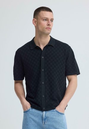 Jeune homme portant une chemise à manches courtes noire à boutons avec un motif à carreaux subtil et un jean bleu clair, les mains dans les poches, regardant vers la droite.