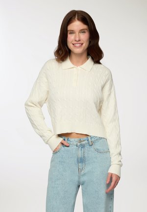 Maglione cropped a maglia grossa color bianco sporco con colletto polo. Presenta polsini e orlo a coste, abbinato a jeans a vita alta di colore blu chiaro.