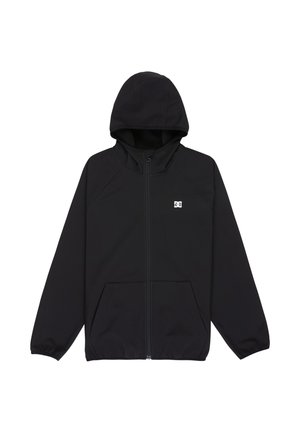 Lachend kind met blond haar, gekleed in een zwarte hoodie, grijze jeans, blauw-witte sneakers en een sjaal met patroon, staand tegen een effen achtergrond.