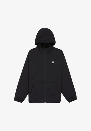 Lächelndes Kind mit blondem Haar, das einen schwarzen Hoodie, graue Jeans, blau-weiße Sneakers und einen gemusterten Schal trägt, steht vor einem schlichten Hintergrund.