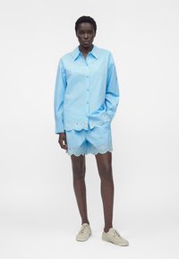 Vrouw draagt lichtblauw overhemd met knopen en bijpassende korte broek met gegolfde oogdetail, gecombineerd met beige sneakers, staand tegen een witte achtergrond.