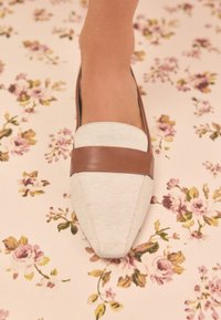 Chaussure plate à bout pointu en tissu texturé blanc avec une bande d'accent en cuir marron sur l'avant, sur un fond à motif floral.