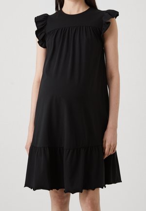 Femme portant une robe noire sans manches avec des épaules à volants et un ourlet évasé sur un fond clair uni.