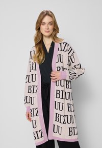 Kobieta z długimi blond włosami, ubrana w czarny strój i jasnoróżowy kardigan z czarnym napisem i lawendowymi wykończeniami, stojąca na szarym tle.