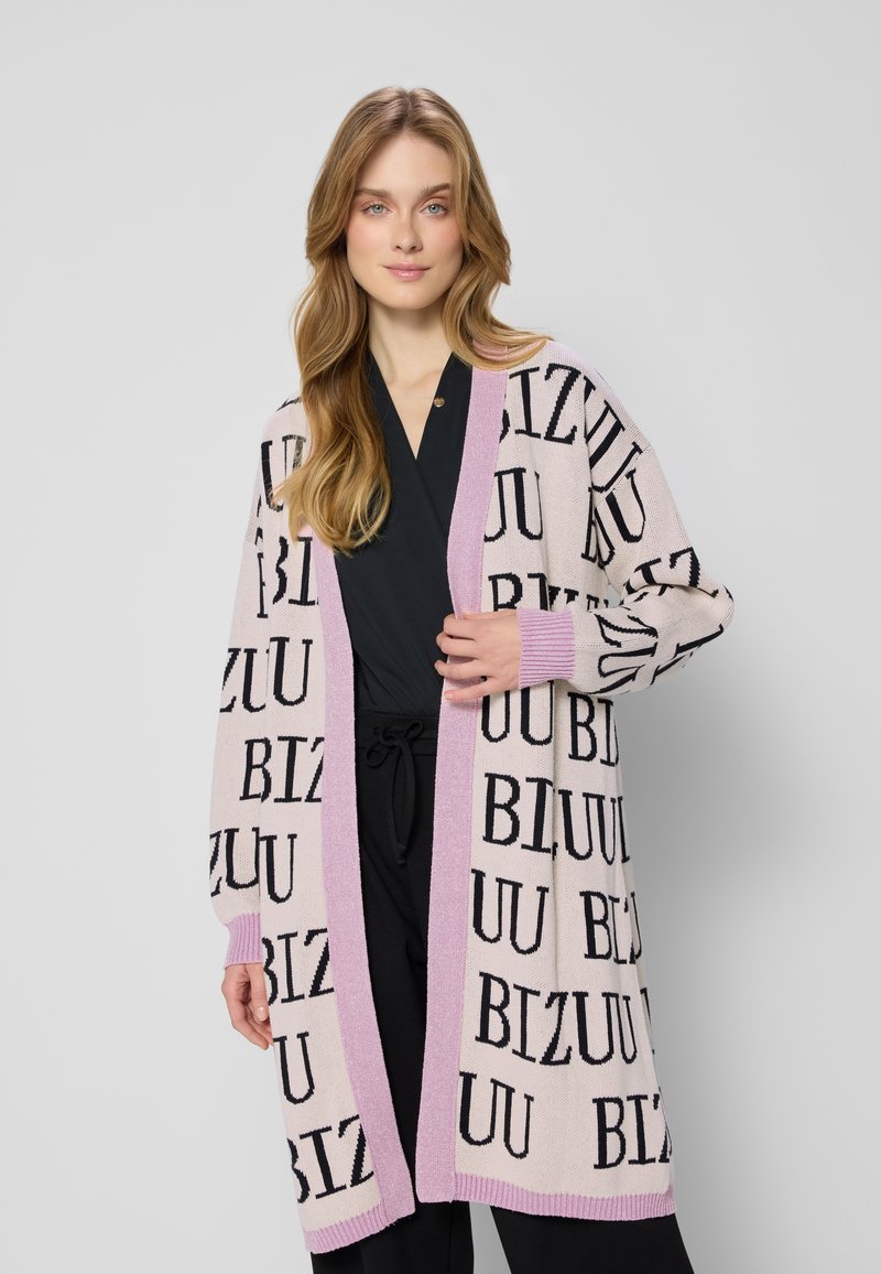 Kobieta z długimi blond włosami, ubrana w czarny strój i jasnoróżowy kardigan z czarnym napisem i lawendowymi wykończeniami, stojąca na szarym tle.