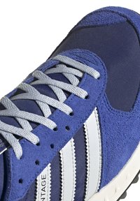 Kék sportcipő, amely velúr és hálós anyagok keverékéből készült. Fehér csíkokkal és szürke fűzőkkel. Textúrázott felsőrésszel és párnázott talppal rendelkezik.