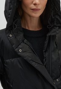 Zwarte gewatteerde jas met een hoge kraag en capuchon, voorzien van een ritsluiting en metalen details. Getextureerde donkere trui eronder.