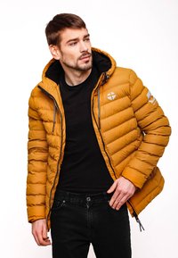 Pufová bunda v horčicovo žltej farbe, s predným zipsom, kapucňou a horizontálnym prešívaním. Pod ňou čierna mikina a tmavé džínsy.