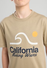 T-shirt in cotone beige con una grafica di onde bianche e testo blu "California Making Waves", con un sole giallo sopra l'onda.