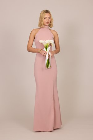 Frau in langem, ärmellosem rosa Kleid, die weiße Calla-Lilien hält, die mit weißem Band vor schlichtem beigem Hintergrund gebunden sind.