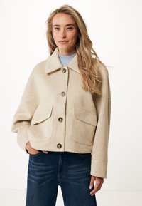 Bežová vlněná bunda se skládacím límcem, velkými předními kapsami a zapínáním na knoflíky. Spárována s tmavě modrými džíny s jemnou texturou.