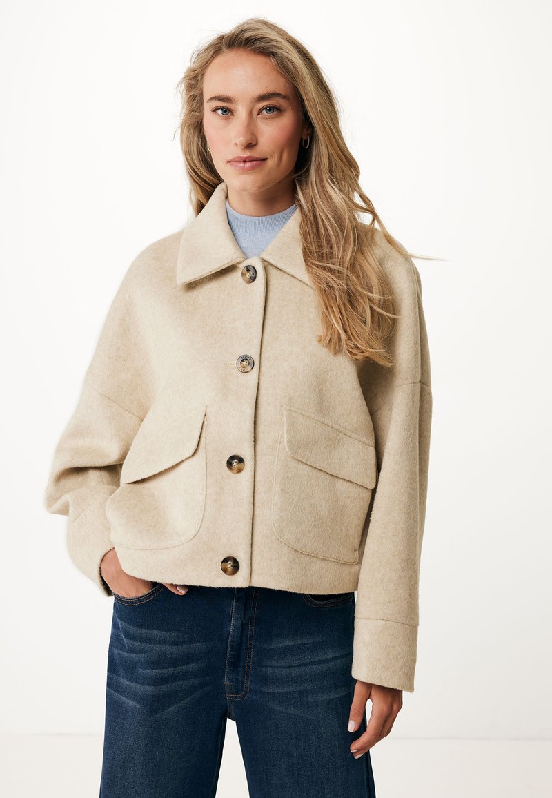 Bežová vlněná bunda se skládacím límcem, velkými předními kapsami a zapínáním na knoflíky. Spárována s tmavě modrými džíny s jemnou texturou.